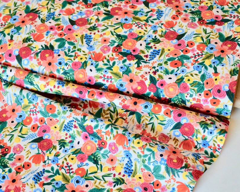 May include: Floral fabric with a dense pattern of colorful flowers. The design features red, pink, orange, yellow, and blue blooms with green leaves on a white background. The fabric is folded, showing the pattern's repeat.