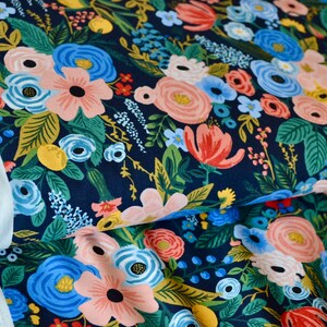 Puede incluir: Primer plano de tela floral con fondo azul marino oscuro. La tela presenta un denso patr&oacute;n de flores coloridas en tonos rosa, azul, rojo y amarillo, con follaje verde. Se ve un borde de tela blanca con el texto "RIFLE PAPER".