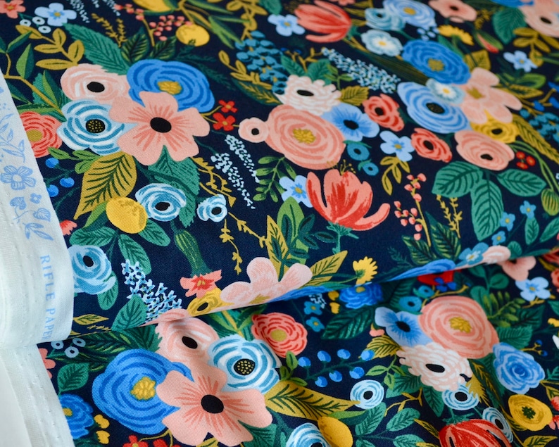 Puede incluir: Primer plano de tela floral con fondo azul marino oscuro. La tela presenta un denso patr&oacute;n de flores coloridas en tonos rosa, azul, rojo y amarillo, con follaje verde. Se ve un borde de tela blanca con el texto "RIFLE PAPER".