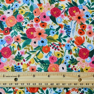 May include: Floral fabric with a white background, featuring a dense pattern of colorful flowers. The flowers are in shades of red, pink, orange, blue, and yellow, with green leaves. A wooden ruler with inch measurements is placed across the fabric.