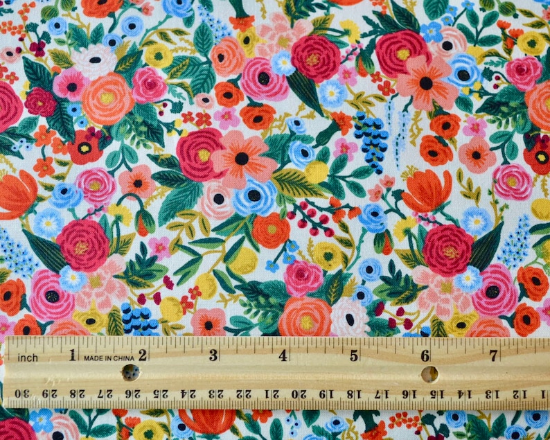 May include: Floral fabric with a white background, featuring a dense pattern of colorful flowers. The flowers are in shades of red, pink, orange, blue, and yellow, with green leaves. A wooden ruler with inch measurements is placed across the fabric.