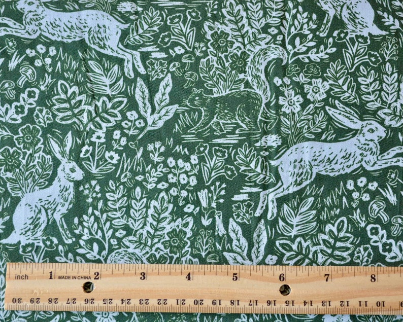 Puede incluir: Tela verde con un estampado blanco de bosque con conejos, ardillas, flores y follaje. El dise&ntilde;o se repite en la tela. Una regla con medidas en pulgadas est&aacute; en la parte inferior de la imagen.