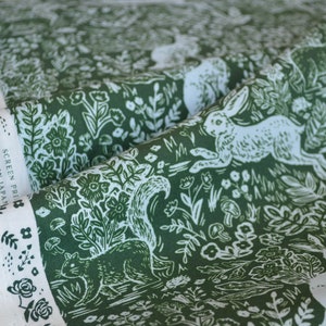 Puede incluir: Tela verde con un dise&ntilde;o de bosque blanco que presenta conejos, ardillas, flores y setas. La tela tiene un borde blanco con el texto "SCREEN PRINT IN JAPAN". La tela est&aacute; doblada, mostrando el patr&oacute;n y el borde.