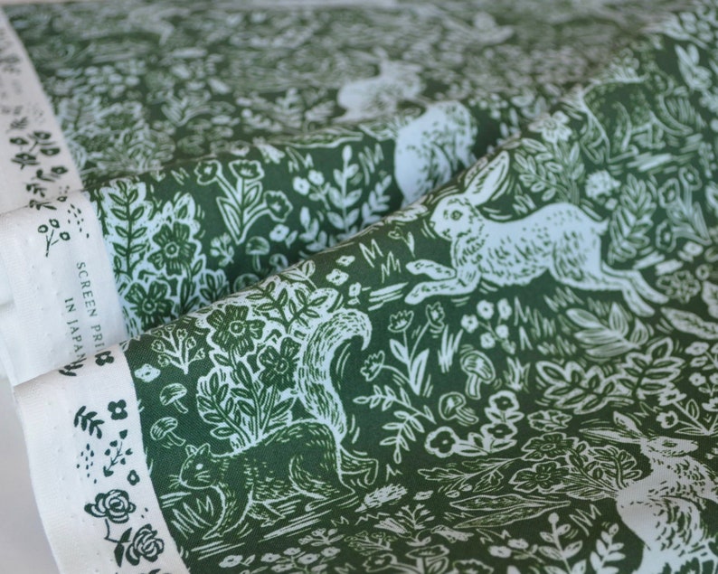 Puede incluir: Tela verde con un dise&ntilde;o de bosque blanco que presenta conejos, ardillas, flores y setas. La tela tiene un borde blanco con el texto "SCREEN PRINT IN JAPAN". La tela est&aacute; doblada, mostrando el patr&oacute;n y el borde.