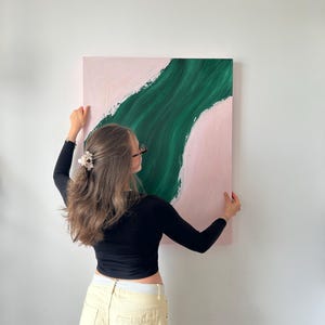 Könnte beinhalten: Abstraktes Kunstwerk mit einer grünen, fließenden Form auf rosa Hintergrund. Die quadratische Leinwand wird vor einer weißen Wand präsentiert. Das Gemälde hat einen modernen, minimalistischen Stil, mit Fokus auf Farbe und Form. Es ist eine quadratische Leinwand.