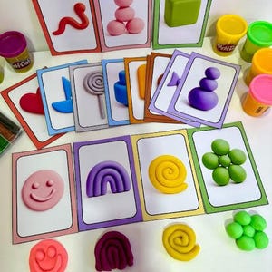 May include: A collection of colorful Play-Doh shapes and cards. The cards feature various shapes like a worm, a smiley face, a rainbow, and a spiral. Small containers of Play-Doh in yellow, green, and pink are also visible.