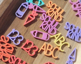 May include: A collection of colorful, plastic school-themed cutouts, including pencils, carrots, numbers, and the letters ABC and the word "SCHU KIND". The cutouts are in shades of orange, purple, pink, yellow, and teal, arranged on a wooden surface.