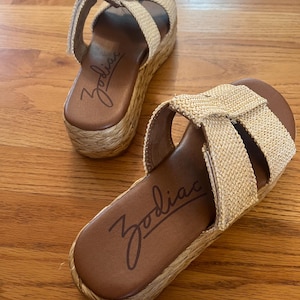 May include: A pair of Zodiac platform sandals. The sandals have woven beige straps and a brown footbed with the Zodiac logo. The platform sole is wrapped in a natural fiber material. The sandals are displayed on a wooden surface.