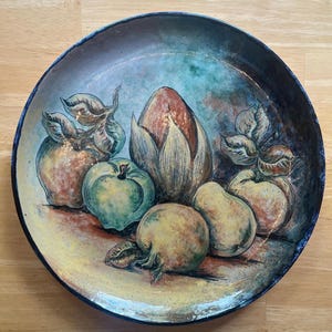 May include: A hand-painted ceramic plate featuring a still life of fruit. The plate has a dark blue rim and a yellow interior. The fruit includes apples, pears, and a central artichoke-like form, rendered in shades of green, yellow, and brown.