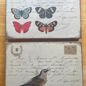 May include: Two decorative wooden panels with a vintage aesthetic. One panel features four butterfly illustrations in shades of blue, red, and orange. The other panel showcases a bird perched on a branch. Both panels have script and stamps.