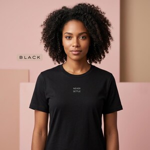 Może przedstawiać: Czarny t-shirt z napisem "NEVER SETTLE" małymi, białymi literami. Słowo "BLACK" znajduje się w beżowym prostokącie w lewym górnym rogu. Osoba nosząca koszulkę ma ciemne, kręcone włosy.