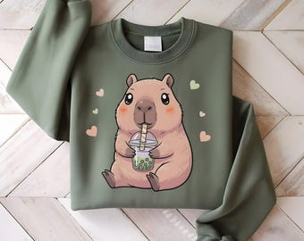 May include: Olive green sweatshirt featuring a cartoon capybara drinking boba tea. The capybara is light brown with pink cheeks and is surrounded by small pink and green hearts. The sweatshirt has long sleeves and a crew neck.