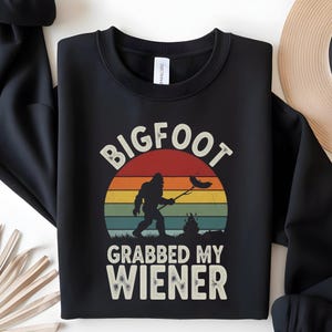 May include: Black sweatshirt with a Bigfoot graphic roasting a sausage over a campfire, and the text "BIGFOOT GRABBED MY WIENER". The design features a sunset with red, orange, yellow, green, and blue stripes.
