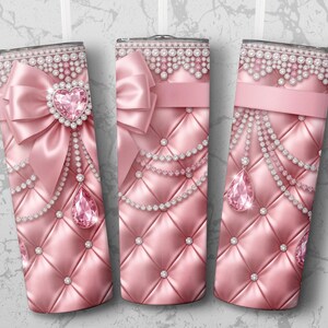 May include: Three pink tumblers with a quilted pattern, adorned with bows, ribbons, and sparkling jewels. Each tumbler features a different design, including heart-shaped gems and dangling teardrop pendants. The tops are embellished with a pearl-like trim.