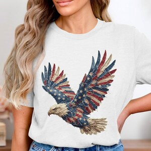 Può includere: T-shirt bianca con una grafica dettagliata di un'aquila calva americana con le ali spiegate. Le ali dell'aquila sono disegnate con le stelle e le strisce della bandiera americana. Un simbolo patriottico.