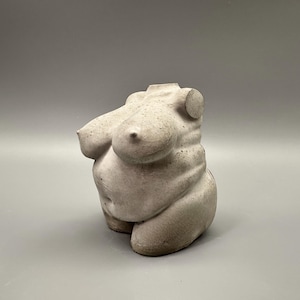 May include: A small, off-white sculpture of a torso. The sculpture depicts a curvaceous form with prominent breasts and a rounded abdomen. The piece is made of a textured material, possibly concrete or plaster, and sits on a plain gray background.
