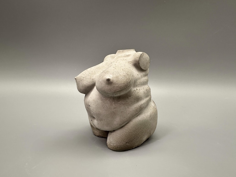 May include: A small, off-white sculpture of a torso. The sculpture depicts a curvaceous form with prominent breasts and a rounded abdomen. The piece is made of a textured material, possibly concrete or plaster, and sits on a plain gray background.