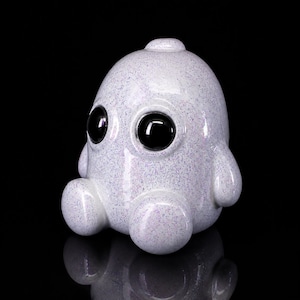 Könnte beinhalten: Eine kleine, abgerundete Figur mit einer glatten, irisierend weißen Oberfläche, die mit feinem, mehrfarbigem Glitzer bedeckt ist. Das Objekt hat zwei große, glänzend schwarze, kreisförmige Augen auf der Vorderseite sowie zwei kleine Arme und Beine.