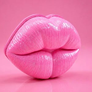 May include: A large, glossy, pink sculpture of a pair of lips. The lips are detailed with vertical lines and have a smooth, reflective surface. The background is a solid pink color.