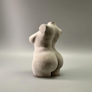 May include: A light-colored, sculpted torso figurine. The sculpture depicts the upper body of a person, with detailed curves and form. The figurine is made of a solid, possibly stone-like material, and is set against a neutral background.