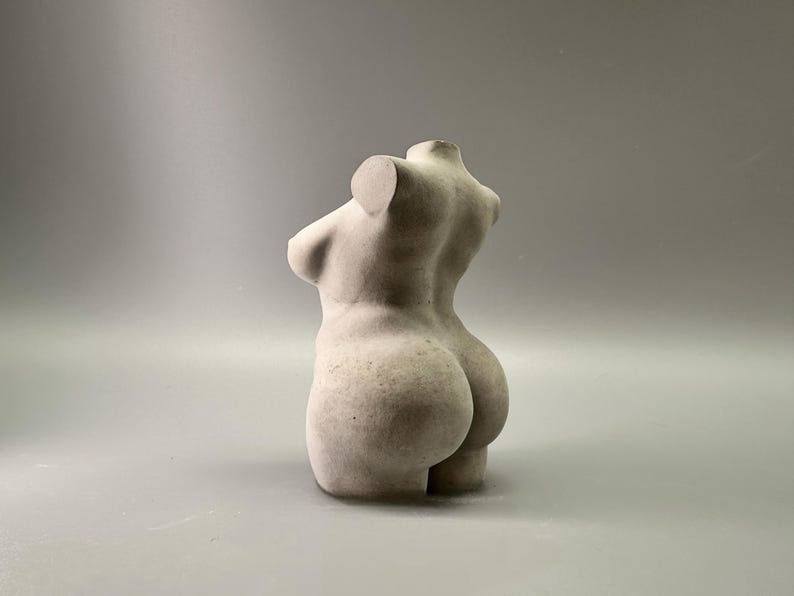 May include: A light-colored, sculpted torso figurine. The sculpture depicts the upper body of a person, with detailed curves and form. The figurine is made of a solid, possibly stone-like material, and is set against a neutral background.
