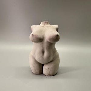 May include: A headless, off-white sculpture of a female torso. The sculpture has a rounded belly, full breasts, and defined legs. The overall form is smooth with subtle details, and it stands on a solid base against a gray background.