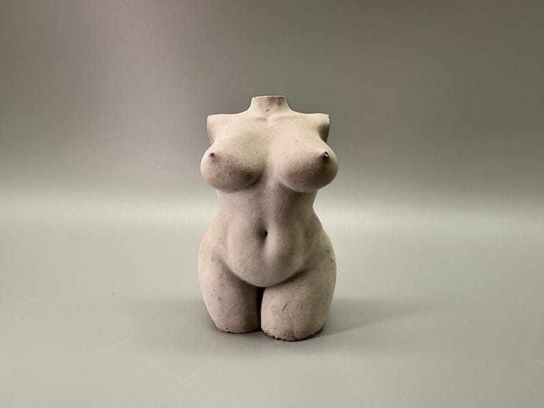 May include: A headless, off-white sculpture of a female torso. The sculpture has a rounded belly, full breasts, and defined legs. The overall form is smooth with subtle details, and it stands on a solid base against a gray background.