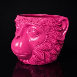 May include: A bright pink monkey-shaped planter or vessel with a textured, fur-like surface. The monkey face features closed eyes, a prominent snout, and a small, distinct red heart shape located on the right cheek area against the vibrant pink.
