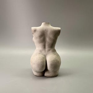 May include: A light-colored, sculpted torso figurine. The back of the figure is visible, showcasing the form's curves and details. The sculpture is a neutral color, with a matte finish, and sits against a plain background.