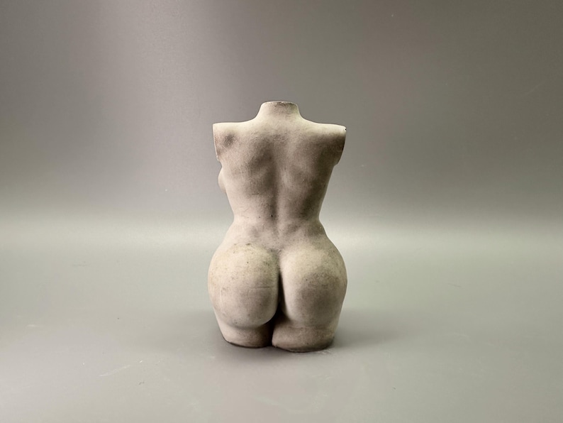 May include: A light-colored, sculpted torso figurine. The back of the figure is visible, showcasing the form's curves and details. The sculpture is a neutral color, with a matte finish, and sits against a plain background.
