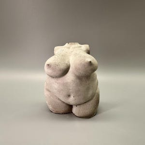 May include: A concrete sculpture of a torso, featuring a rounded belly and prominent breasts. The sculpture is a light gray color and has a textured surface. The artwork is a representation of the female form.