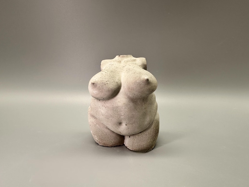May include: A concrete sculpture of a torso, featuring a rounded belly and prominent breasts. The sculpture is a light gray color and has a textured surface. The artwork is a representation of the female form.