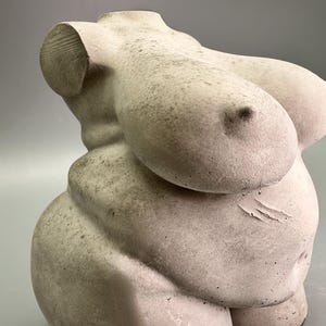 May include: A light-colored, sculpted torso statue. The sculpture depicts a female form with prominent breasts and a rounded abdomen. The statue has a textured surface and is set against a gray background. The sculpture is a decorative art piece.