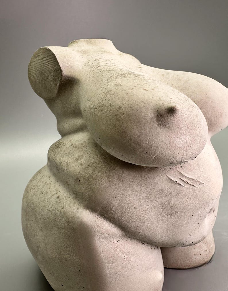 May include: A light-colored, sculpted torso statue. The sculpture depicts a female form with prominent breasts and a rounded abdomen. The statue has a textured surface and is set against a gray background. The sculpture is a decorative art piece.