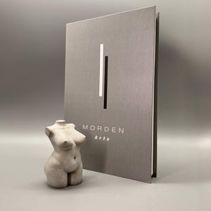 May include: A gray sculpture of a female torso stands next to a gray book with the text "MORDEN Arts". The book has a vertical white and black stripe design. The sculpture is a light gray color and has a smooth texture.