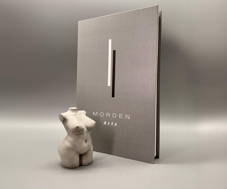 May include: A gray sculpture of a female torso stands next to a gray book with the text "MORDEN Arts". The book has a vertical white and black stripe design. The sculpture is a light gray color and has a smooth texture.