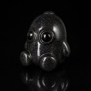 Könnte beinhalten: Eine kleine, abgerundete Figur aus dunklem, glitzerndem Material, das an schwarzen Sandstein erinnert. Das Objekt hat zwei große, glatte, kreisförmige schwarze Augen auf der Vorderseite, einen bauchigen Kopf und zwei kleine, abgerundete Gliedmaßen.