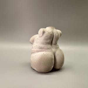 May include: A light gray, sculpted torso figurine. The sculpture depicts the back of a human torso, with the shoulders, back, and buttocks visible. The figurine has a rough, textured surface and is set against a gray background.