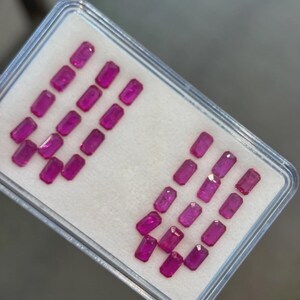 Può includere: Un contenitore di plastica trasparente che contiene numerose pietre preziose rosa vibranti, tagliate a forma rettangolare. Le pietre sono disposte ordinatamente su una superficie bianca, creando un'esposizione visivamente accattivante.
