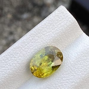 May include: An oval-cut gemstone with a mix of yellow and green hues. The gem is set against a white surface, showcasing its facets and color variations. The stone appears to be a natural gemstone, possibly a tourmaline or similar. The gem is approximately 1 inch in length.