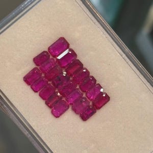 Pode incluir: Uma coleção de pedras preciosas retangulares, facetadas, rosa escuro, dispostas numa caixa de plástico transparente. As pedras têm uma cor vibrante e saturada e um aspeto polido, sugerindo que são rubis ou pedras preciosas semelhantes.