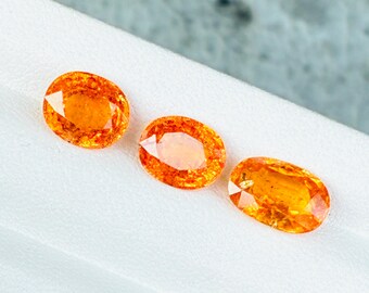 May include: Three oval-cut, orange gemstones are displayed on a white surface. The gems have a faceted appearance, catching the light and showcasing their vibrant color. The gemstones are arranged in a row, each with a unique shape and size.