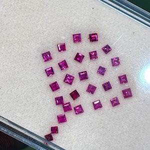 May include: A selection of square-cut, vivid pink gemstones, possibly rubies, are displayed on a white, textured surface. The gems show slight colour variations, with some appearing a deeper crimson. The arrangement suggests a loose grouping of individual stones.
