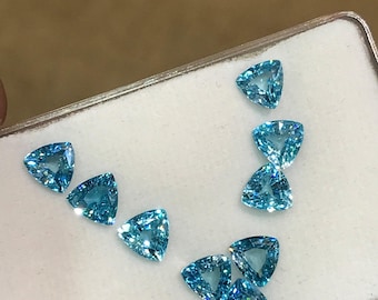 May include: A collection of brilliant, triangular-cut, light blue gemstones. The gems are displayed on a white surface within a rectangular container. The gemstones are faceted, reflecting light and creating a sparkling effect. The background is a warm, neutral tone.