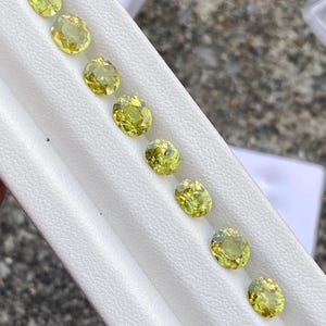 May include: A collection of eight oval-cut, light yellow-green gemstones arranged in a row on a white surface. The gems have a faceted appearance, reflecting light. The background is a blurred outdoor setting.