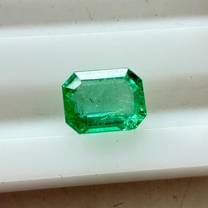 Könnte beinhalten: Ein lebendiger, smaragdgeschliffener grüner Edelstein. Der Stein ist rechteckig mit abgeschrägten Kanten und zeigt einen satten, transparenten grünen Farbton. Die Facetten fangen das Licht ein und unterstreichen die Klarheit und Brillanz des Steins. Der Edelstein liegt auf weißem Hintergrund.