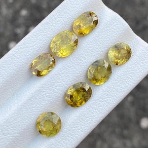 May include: A collection of oval-cut, yellow gemstones. The gems are arranged on a white surface, showcasing their faceted surfaces and golden hues. The gemstones vary slightly in color, ranging from light to deep yellow.
