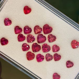 May include: A collection of heart-shaped gemstones in varying shades of red and pink. The gems are displayed on a white surface within a clear, rectangular container. The gemstones are faceted and reflect light.