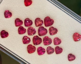 May include: A selection of heart-shaped gemstones in shades of red and pink. The gems are presented on a white surface within a clear, rectangular container. The gemstones are faceted and reflect light.