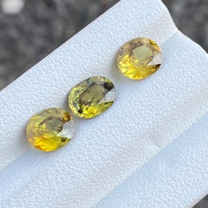 May include: Three oval-cut gemstones with a yellow-green hue are displayed on a white surface. The gems have a faceted cut, reflecting light. The gemstones are likely for jewelry making or collecting.